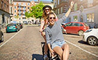 Grinning pretty young woman waving at the camera as she rides on the handlebars of a friends bicycle down a cobbled urban street